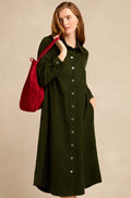 A woman stands against a plain beige background, wearing the Forest Pinwale Cord Shirt Dress—a long-sleeve, button-down, dark green corduroy dress with a breast pocket—while carrying a red shoulder bag.