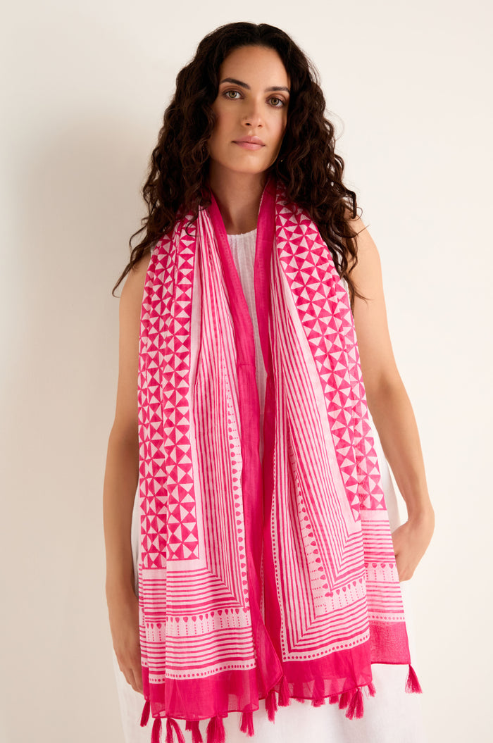 A woman with long curly hair, wearing a white dress and the Fuchsia Tassel Cotton Scarf—with geometric pink print and tassel edging—stands against a plain light background.