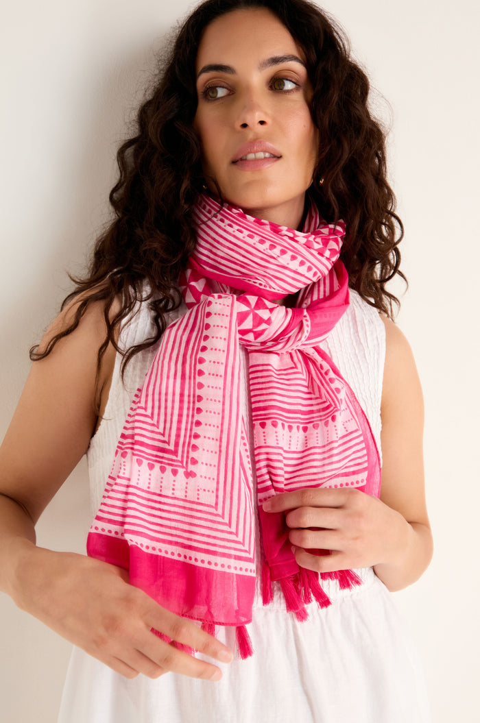 A woman with long curly hair wearing a white sleeveless top and the Fuchsia Tassel Cotton Scarf, featuring a pink geometric print and tassel edging, stands against a plain light-colored background.