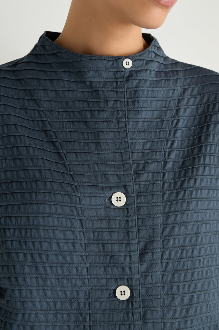 Close-up of a person wearing the Funnel Neck Stone Blue Cotton Jacket featuring a dark textured fabric, stand collar, white buttons, and boxy silhouette; the person’s face is partially out of frame.