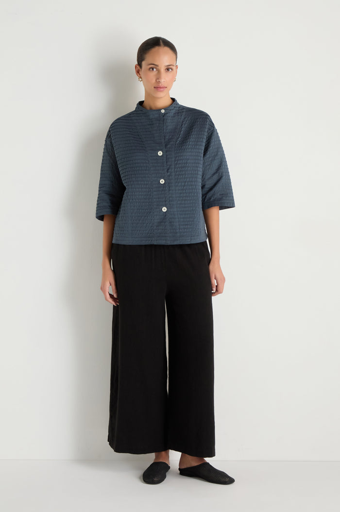 A person stands against a plain white wall wearing the Funnel Neck Stone Blue Cotton Jacket with a boxy silhouette, paired with black wide-leg pants and black shoes.