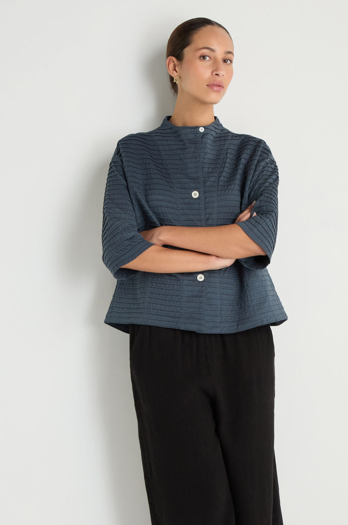 A person stands against a white wall, arms crossed, wearing a Funnel Neck Stone Blue Cotton Jacket with a boxy silhouette and three-quarter sleeves, paired with loose black pants.