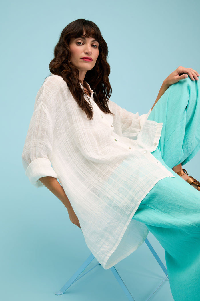 A person with long brown hair sits on a blue chair, wearing the Gauzy Linen Relaxed Shirt and turquoise pants against a light blue background.