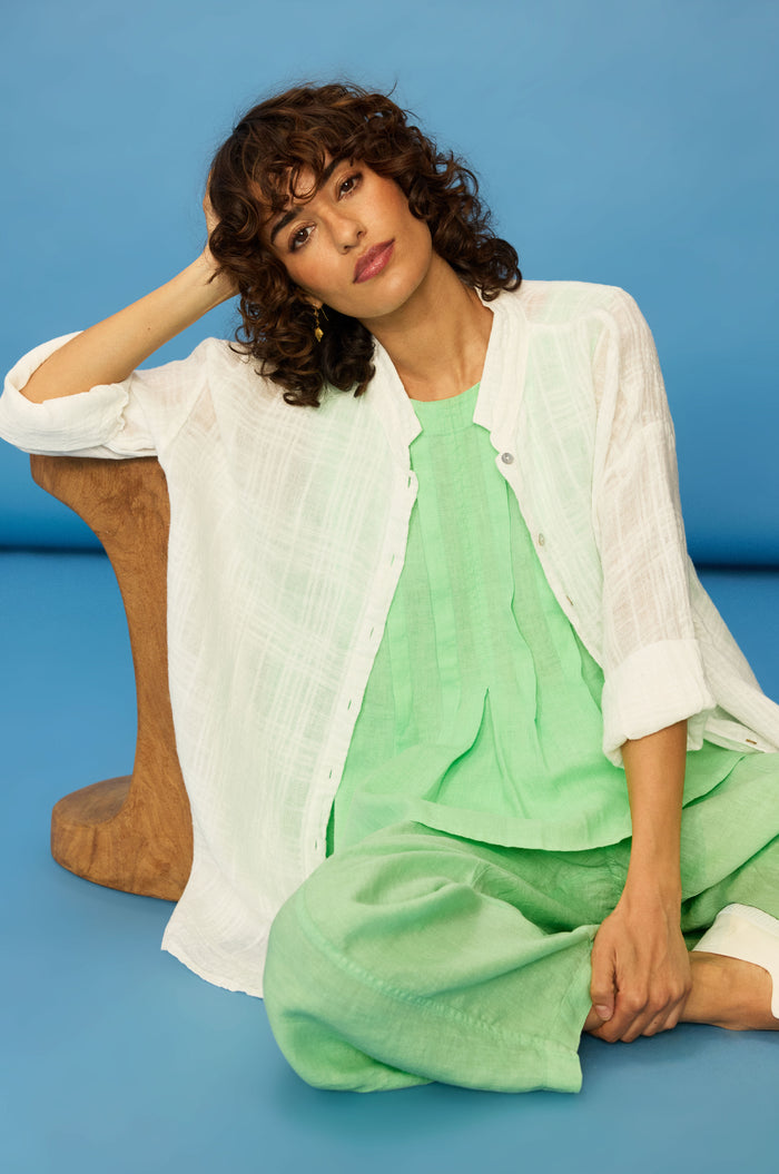 A woman with curly hair sits against a blue background, wearing the Gauzy Linen Relaxed Shirt over a mint green outfit, with one arm resting on a wooden object.