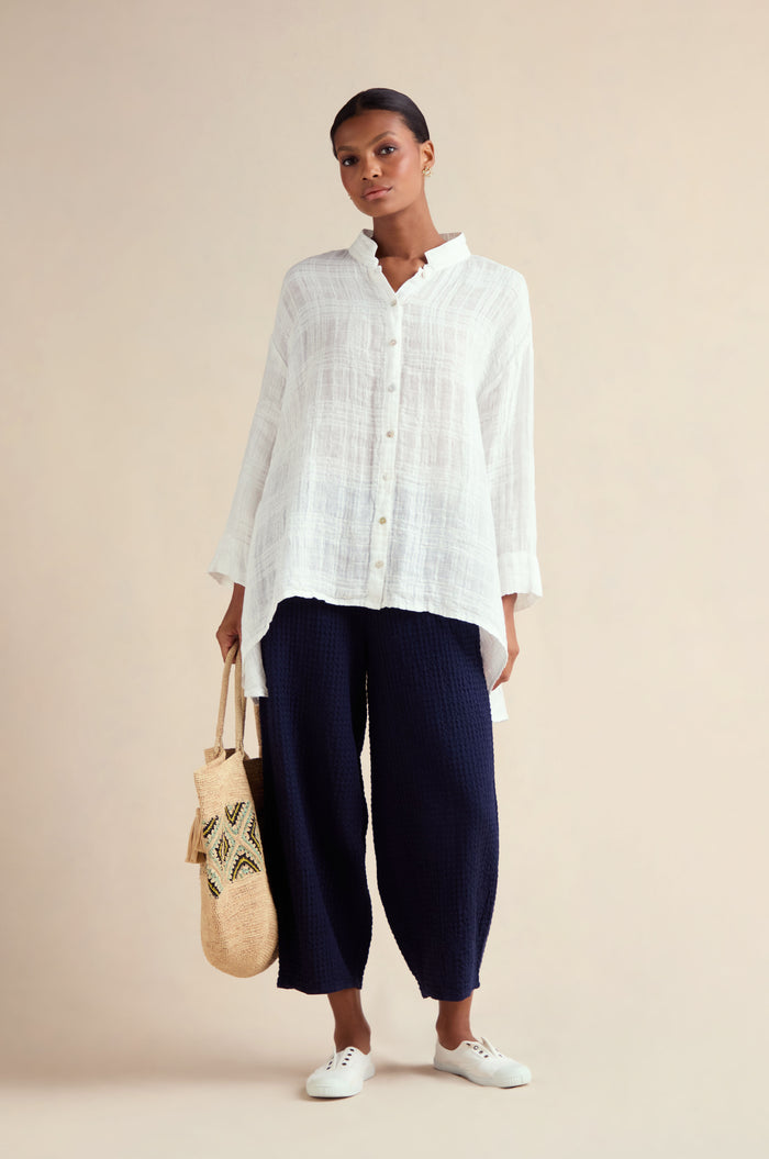 A woman stands against a plain background wearing the Gauzy Linen Relaxed Shirt, navy textured wide-leg pants, white sneakers, and holding a woven tote bag.
