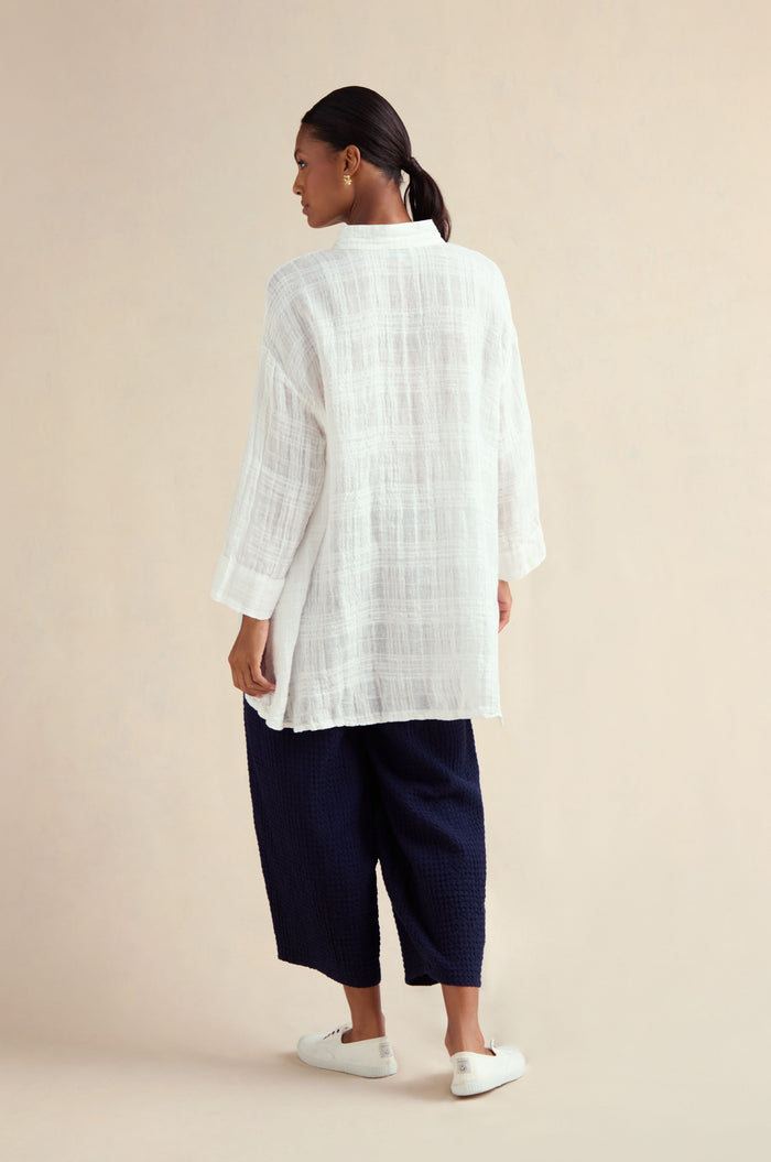 A woman stands with her back to the camera, wearing the Gauzy Linen Relaxed Shirt, dark wide-leg pants, and white slip-on shoes against a neutral background.