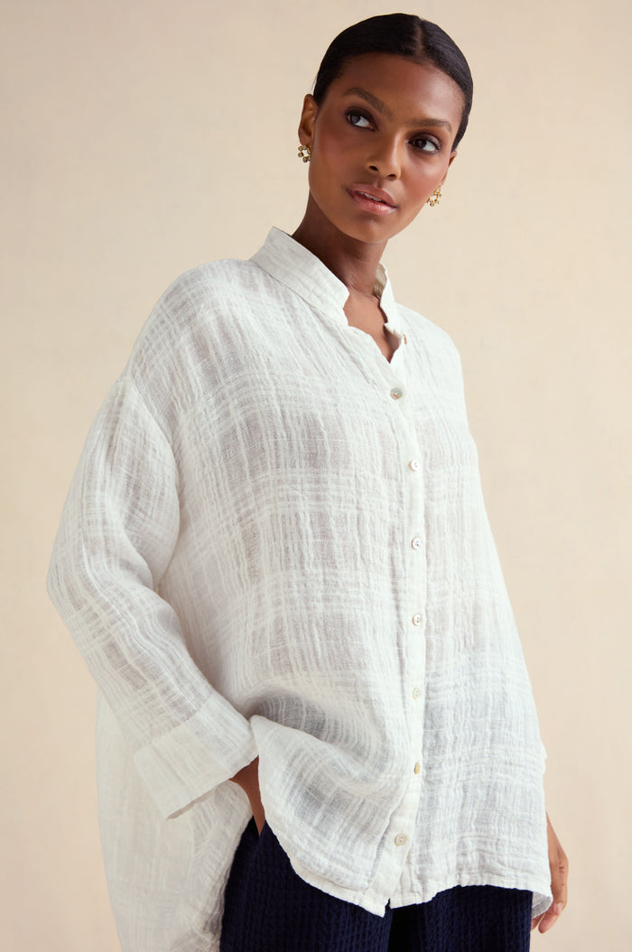 Woman wearing the Gauzy Linen Relaxed Shirt in white with a subtle woven check texture, paired with dark pants and gold earrings, standing against a light background.