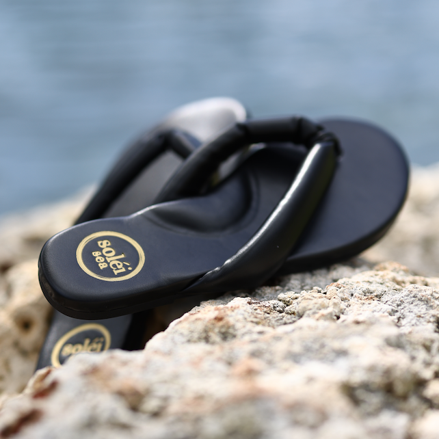 Black rubber flip-flops with round gold-tone logo on the straps, resting on a textured coastal rock.