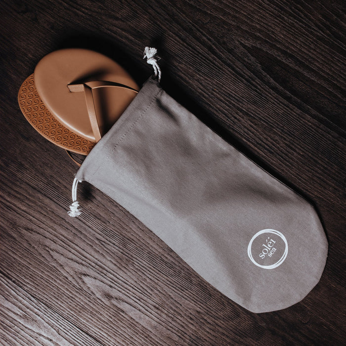 Gray drawstring fabric pouch with white circular logo, resting on dark wood with tan thong sandals.
