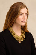 A woman with wavy auburn hair, dressed in a black top, wears the Gold Beaded Collar Necklace—a large, multi-strand accessory—and looks slightly to the right against a neutral background.