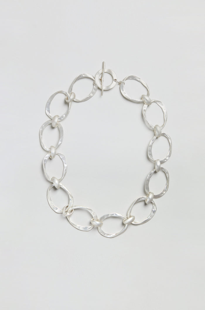 The Hammered Silver Hoop Necklace showcases large, irregular oval links with a hammered finish, joined by smaller round connectors, and is elegantly presented against a plain white background.