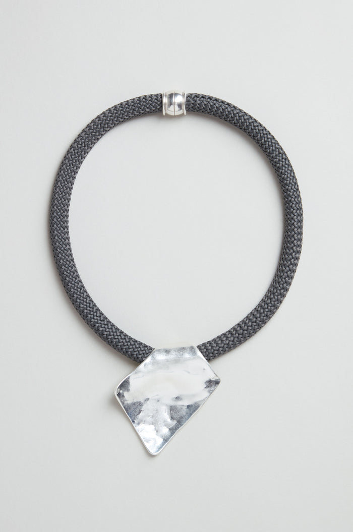 The Hammered Silver Pendant Cord Necklace features a black braided cord, an irregularly-shaped polished hammered silver pendant, and a silver clasp, shown on a light gray background.