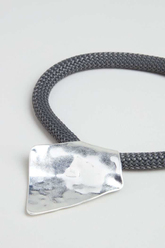 The Hammered Silver Pendant Cord Necklace features a thick, braided dark gray cord with a large, irregularly shaped hammered silver pendant, displayed elegantly on a light background.
