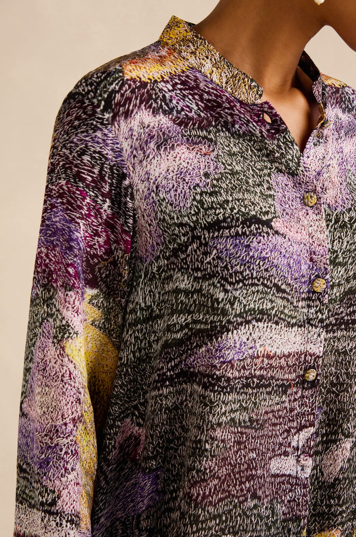 Wearing the Impressionist Print Flared Shirt, a person showcases a collared, button-up design made from a luxurious blend of cupro and viscose, adorned with a textured multicolor pattern in shades of purple, black, yellow, and white.
