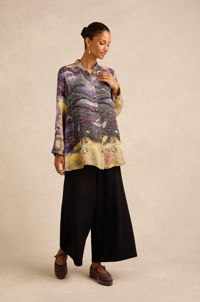 A woman stands wearing the Impressionist Print Flared Shirt, crafted from a luxurious cupro and viscose blend, paired with wide black pants and brown shoes against a neutral background.