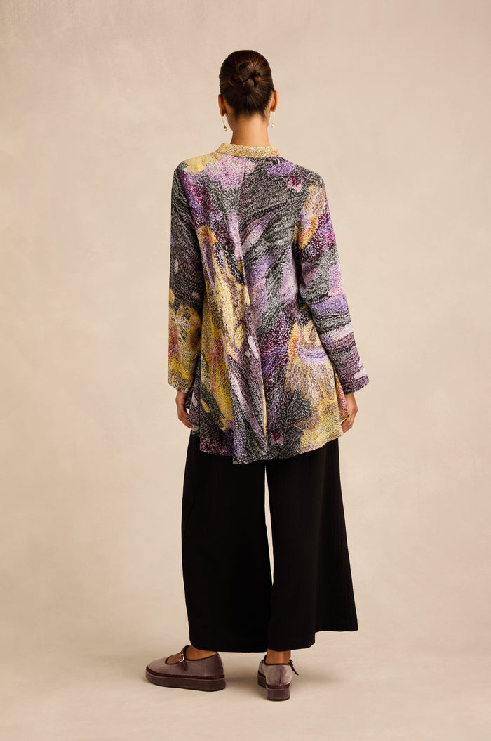 A person stands facing away, wearing the Impressionist Print Flared Shirt—a long-sleeve, multicolored patterned tunic crafted from a luxurious cupro and viscose blend—paired with wide black pants and brown shoes against a neutral background. Hair is styled in a bun.