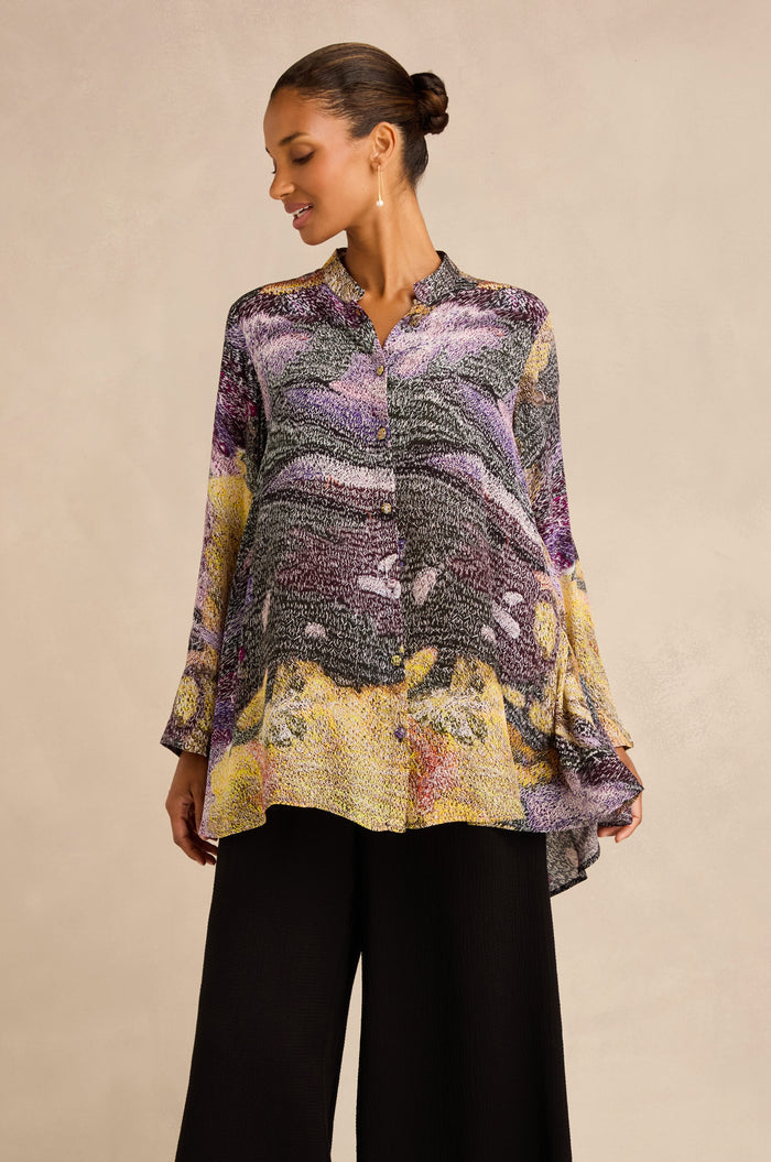A woman models the Impressionist Print Flared Shirt, a loose, long-sleeved blouse crafted from a luxurious blend of cupro and viscose. Showcasing abstract patterns in purple, yellow, and black, this artisan-produced top is paired with wide-leg black pants. She stands against a plain beige background.