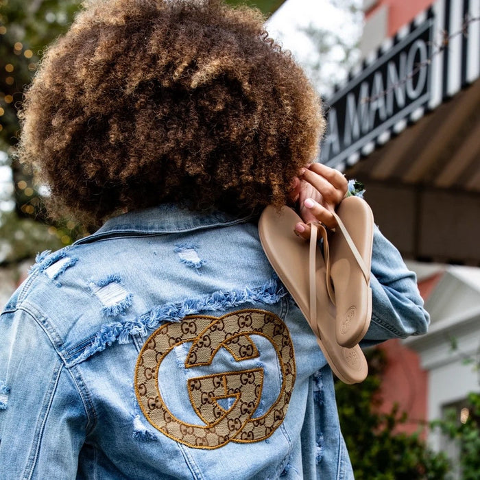 Curly-haired person seen from behind wearing a denim jacket with round embroidered patch holding tan flip-flops