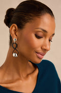 A woman with smooth brown skin and dark hair in a low bun, wearing a teal top and the Infinity Earrings—large, sculptural silver and black earrings handmade in France—looks downward with a slight smile.