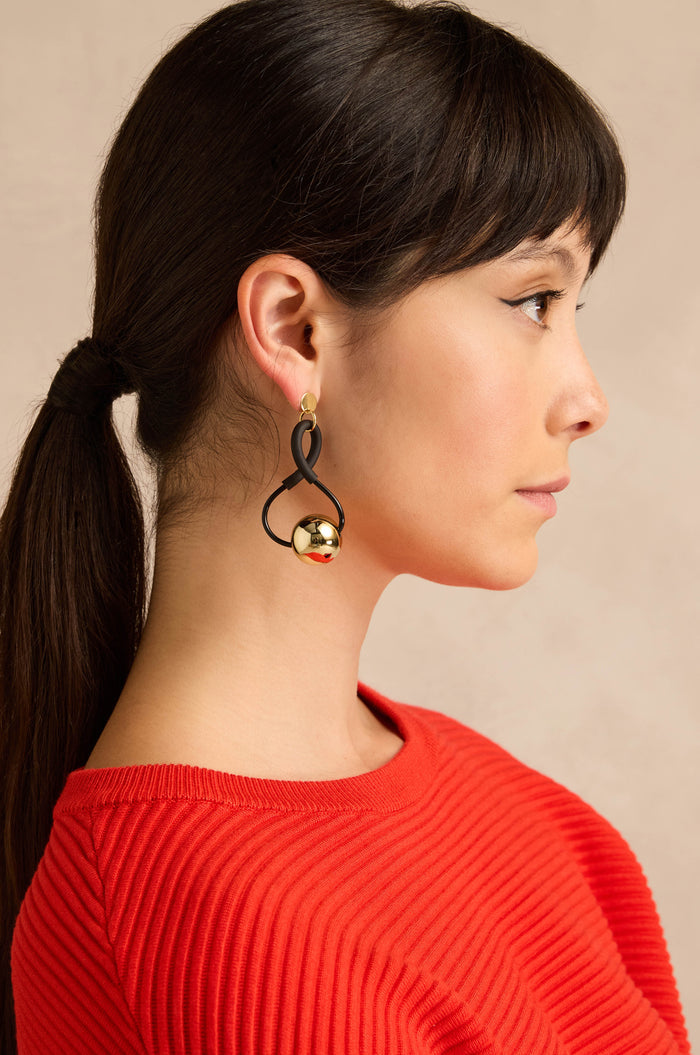 A woman in profile with brown hair styled in a ponytail wears a red ribbed top and the Infinity Earrings—eye-catching black and gold earrings with a sculptural design—set against a beige background.