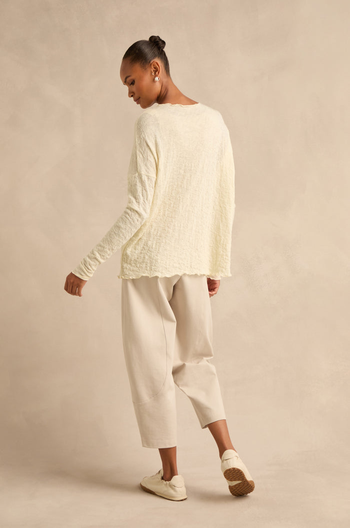 A person stands sideways against a beige studio background, wearing the Ivory Textured Merino Scoop Neck Top, loose light beige pants, and cream slip-on shoes.