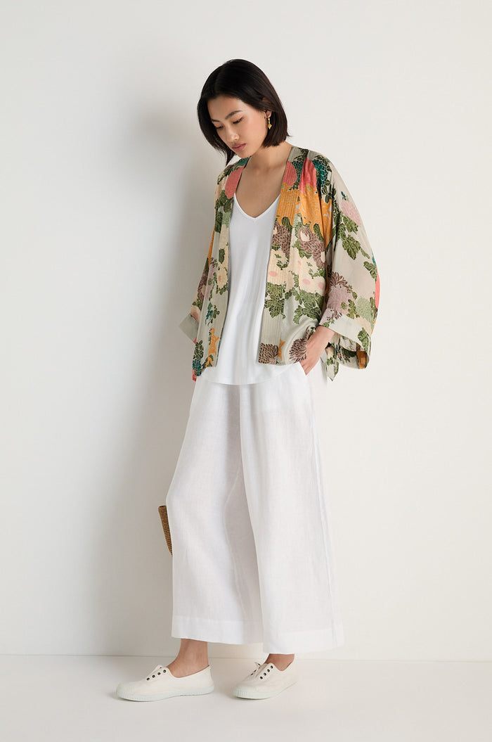 A woman stands against a white wall wearing the Jacquard Kimono—crafted from a soft modal and cupro blend—over a white top, wide-leg white pants, and lace-up shoes, looking downward with one hand in her pocket.