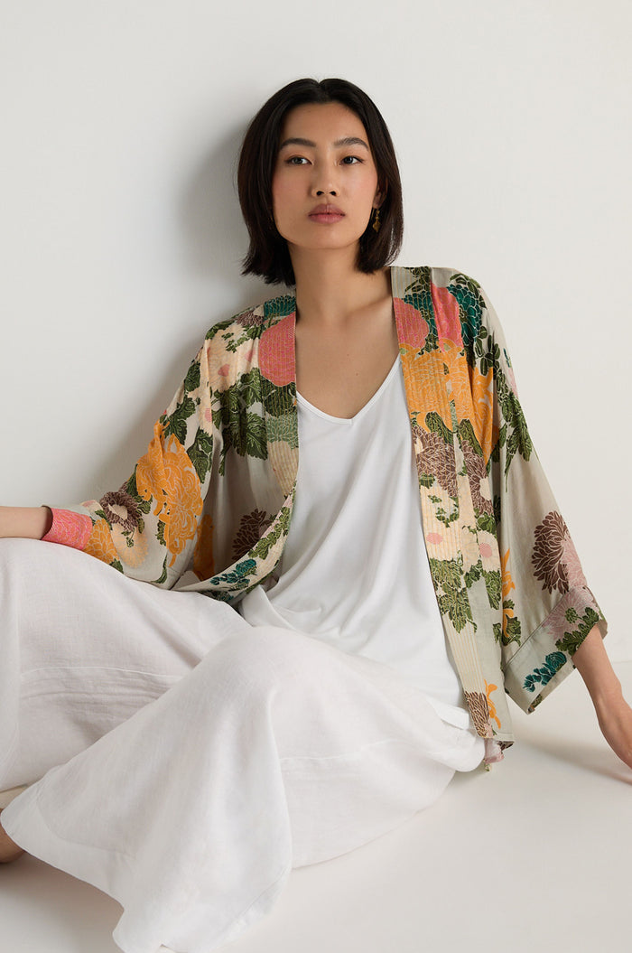 A woman with a bob haircut models the Jacquard Kimono with a floral print over a white top and wide-leg white pants, seated against a light background.
