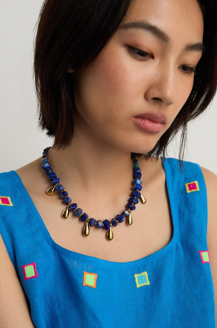 A woman with short dark hair wears a blue dress with small square designs and a Lapis Lazuli Teardrop Necklace featuring blue beads and gold teardrop accents.