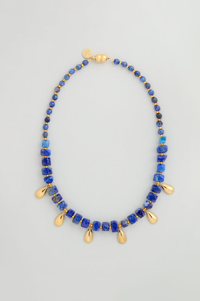 The Lapis Lazuli Teardrop Necklace is handmade in Scotland, featuring blue lapis lazuli stones and gold teardrop pendants, finished with a gold clasp and a small gold tag.