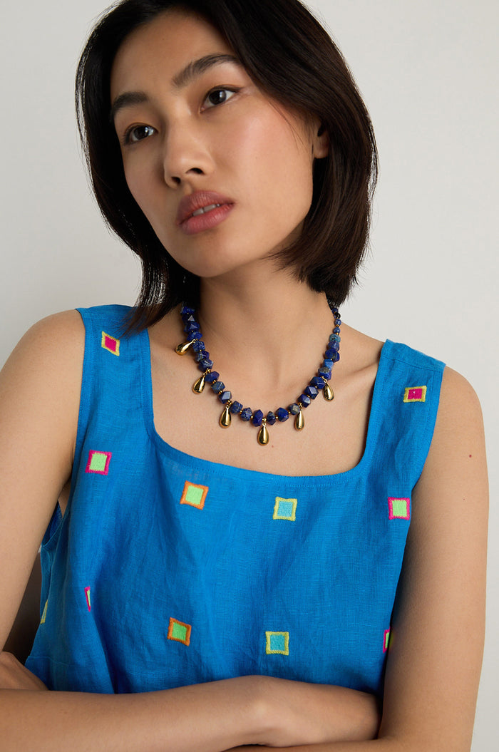 A woman with short dark hair wears a blue sleeveless top with colorful squares and a handmade Lapis Lazuli Teardrop Necklace from Scotland, featuring bold blue and gold beads.