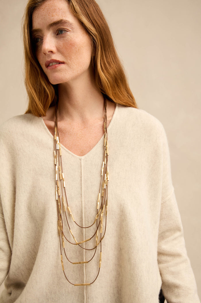 Woman with long auburn hair wearing a cream-colored V-neck sweater and multiple layered gold necklaces, including the striking Leather and Bead Cascade Necklace, looking slightly to the side against a neutral background.