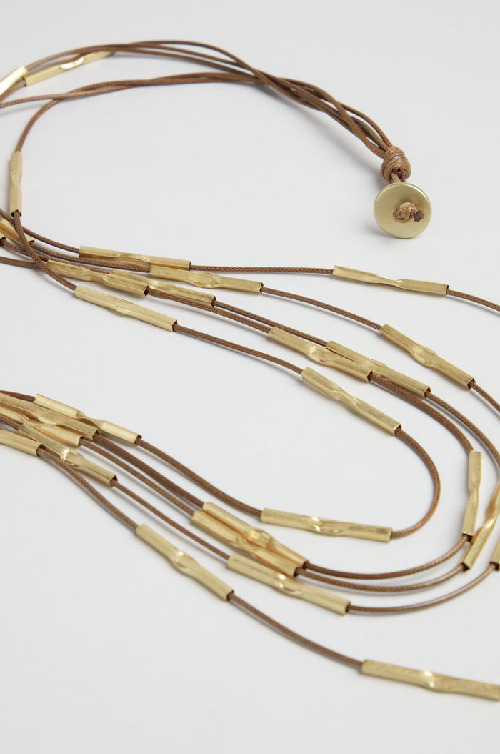 The Leather and Bead Cascade Necklace features long brown leather strands, each adorned with hammered gold cylindrical beads and finished with a round gold-toned button clasp for a striking layered look.