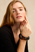 A woman with long red hair, dressed in a black top, poses with her hand near her face to showcase the Leopard Bangle—a wide resin accessory featuring a glossy leopard print finish.