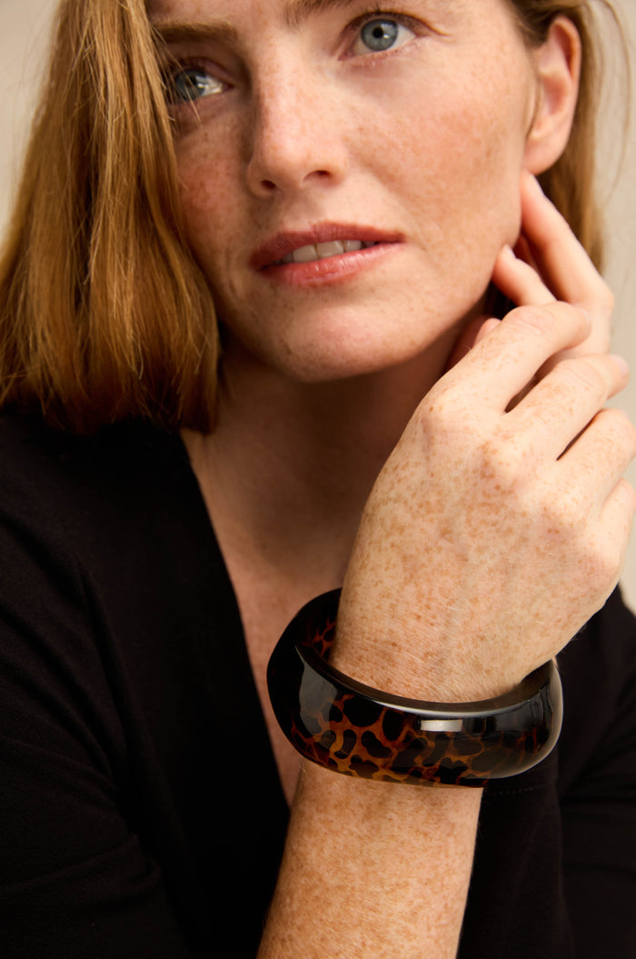 A woman with light skin and freckles, dressed in a black top, rests her hand on her cheek while wearing the glossy-finished Leopard Bangle—a chunky tortoiseshell bracelet.