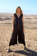 A woman in the Linen Relaxed Jumpsuit and sandals stands on a rocky desert landscape with mountains in the background.