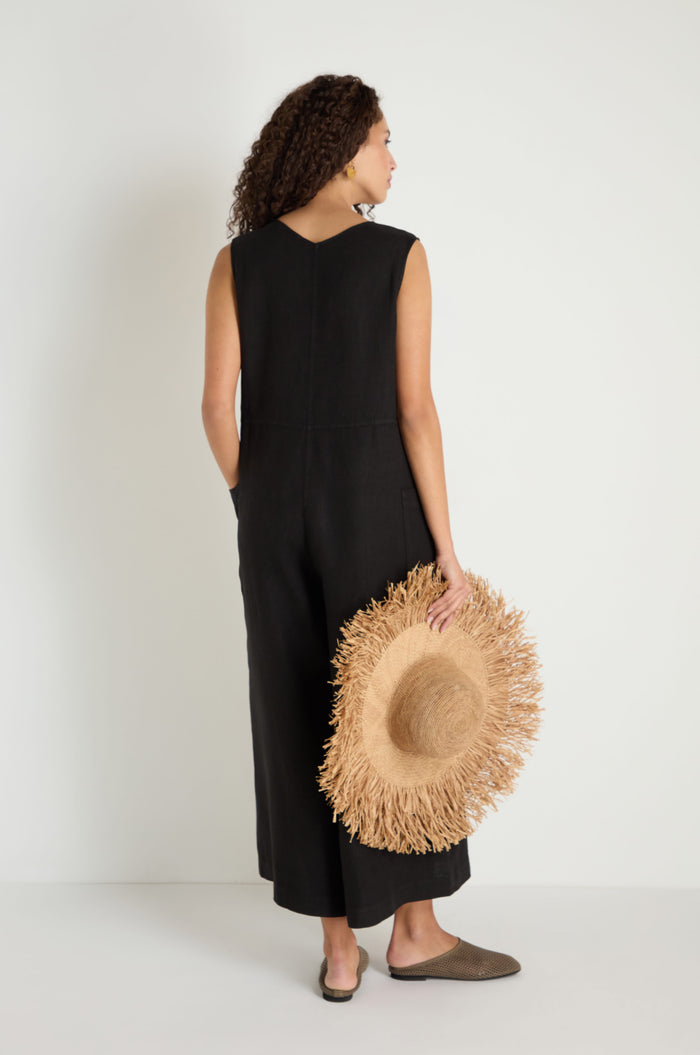 A woman faces away in the Linen Relaxed Jumpsuit, a sleeveless black wide-leg linen piece. She holds a straw hat, her curly hair showing, and wears brown slip-on shoes.