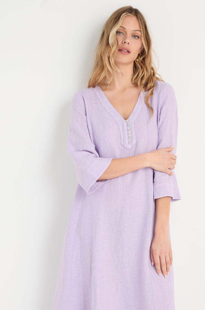 A woman with long blonde hair wears the Linen Seam Pansy Flare Dress—a light purple, V-neck, long-sleeve flare dress with panel seams and decorative buttons—standing against a plain white background.