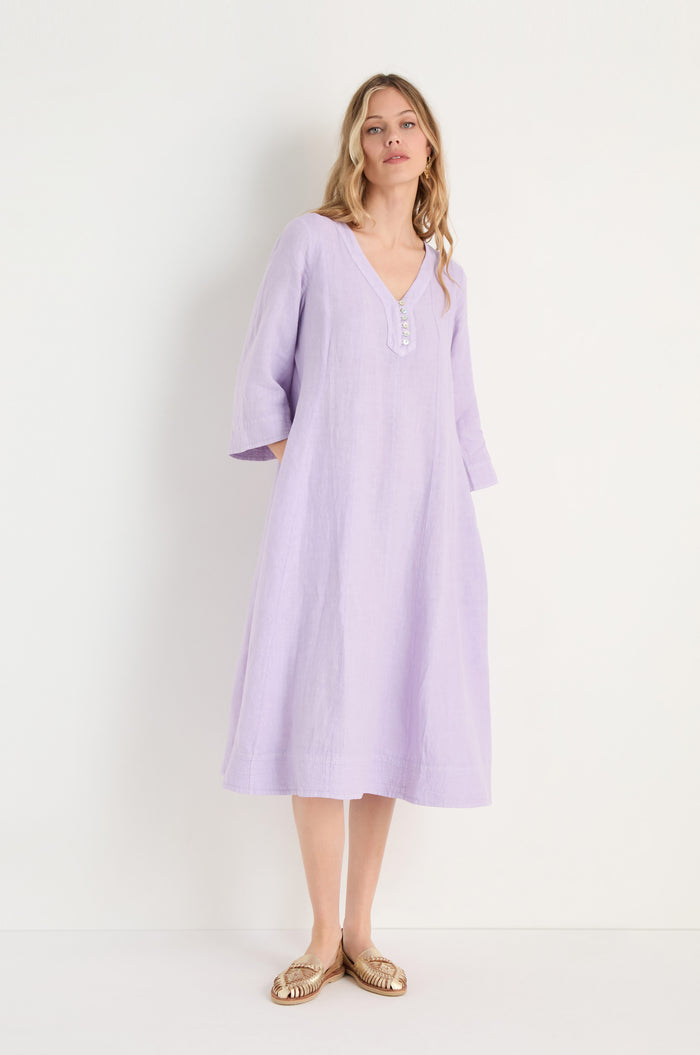 A woman wears the Linen Seam Pansy Flare Dress in light purple, featuring a loose fit, flare silhouette, three-quarter sleeves, and subtle panel seams, standing against a plain white background.