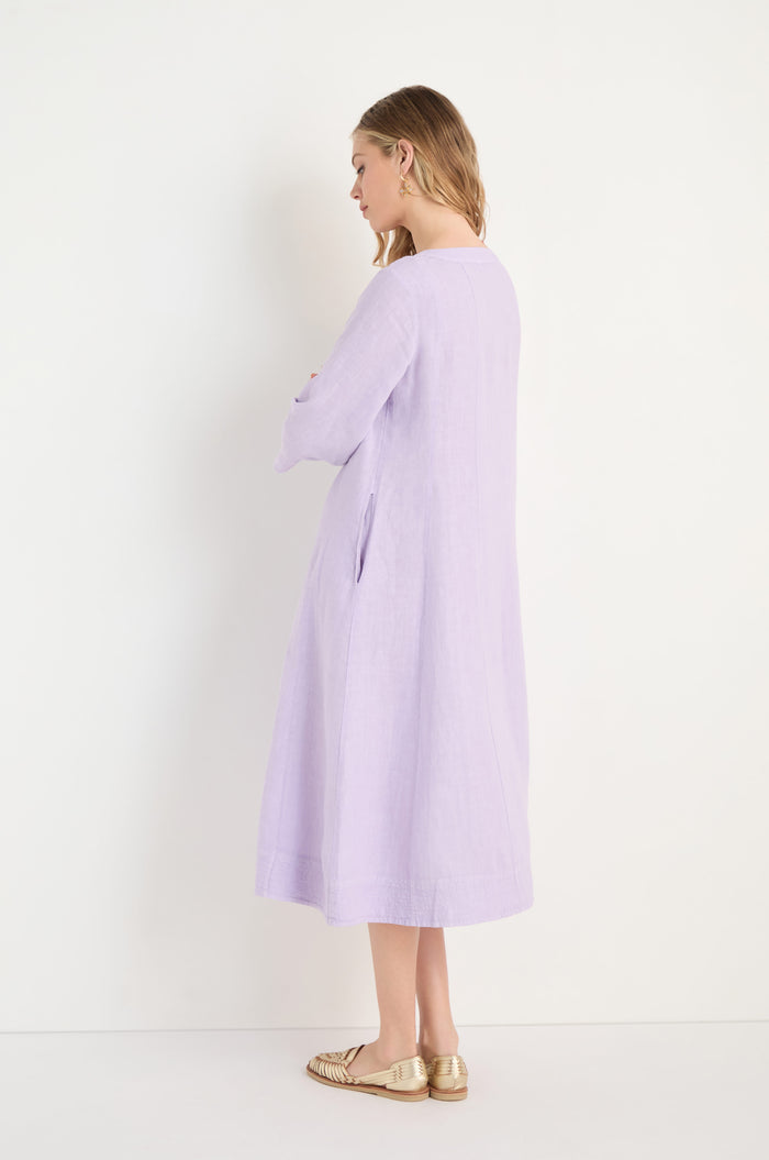 A woman stands sideways with hands crossed, wearing the Linen Seam Pansy Flare Dress in light purple with long sleeves and panel seams, paired with gold flat shoes, set against a plain white background.