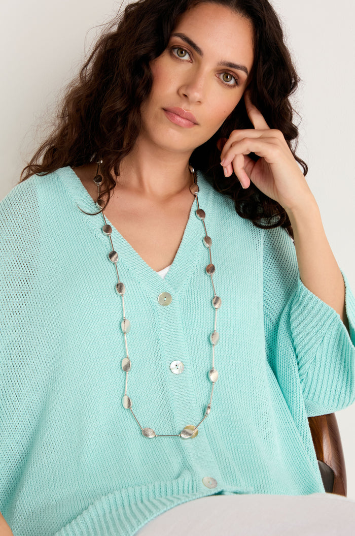 A woman with long curly hair wears a light blue buttoned sweater and the Long Oval Disc Necklace, featuring a metallic finish, as she sits and looks at the camera.