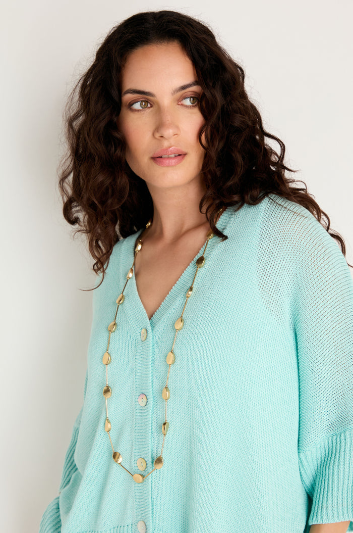 A woman with curly dark hair wears a light blue knit cardigan and the Long Oval Disc Necklace, featuring textured oval discs, as she looks slightly to the side against a plain background.