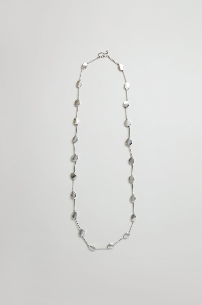 The Long Oval Disc Necklace features textured silver oval discs evenly spaced along a thin chain, shown against a plain light background.