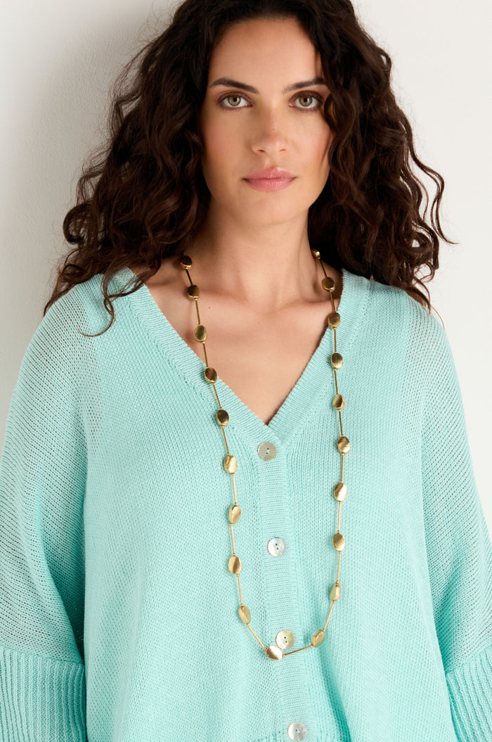 A woman stands against a plain light background, wearing a light blue buttoned sweater and the Long Oval Disc Necklace featuring textured oval discs.