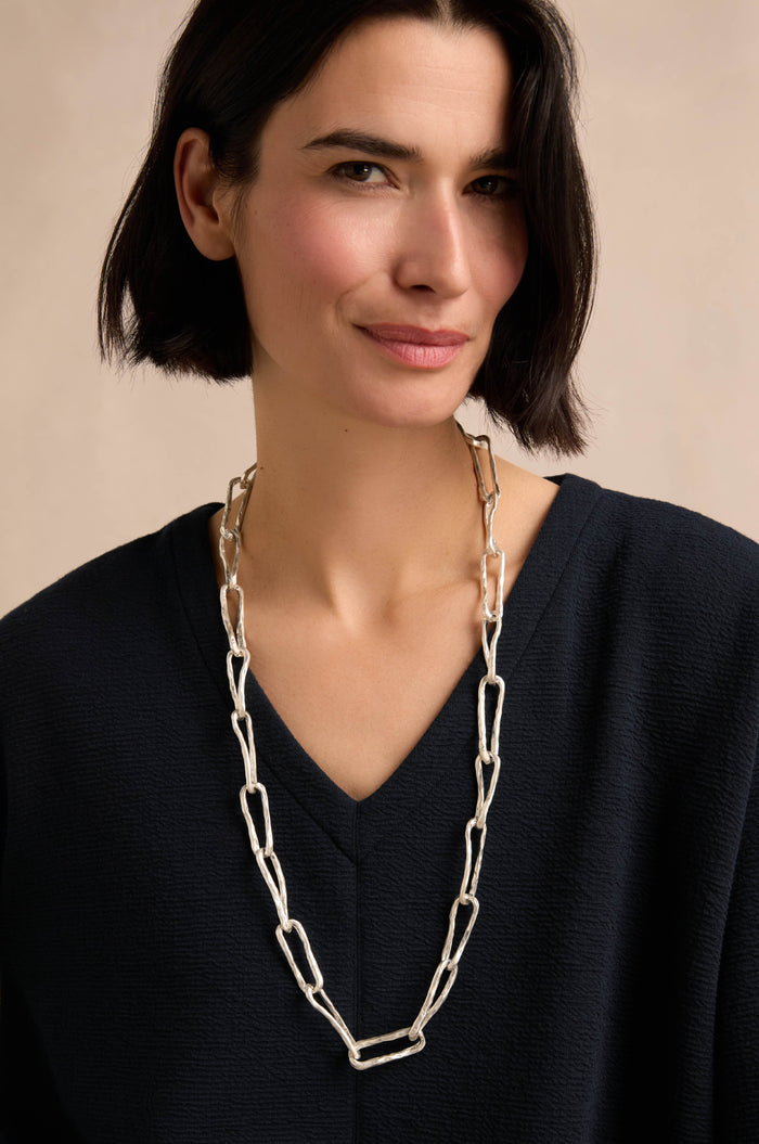 A person with short dark hair wearing a black top and the Long Silver Links Necklace stands against a neutral background.