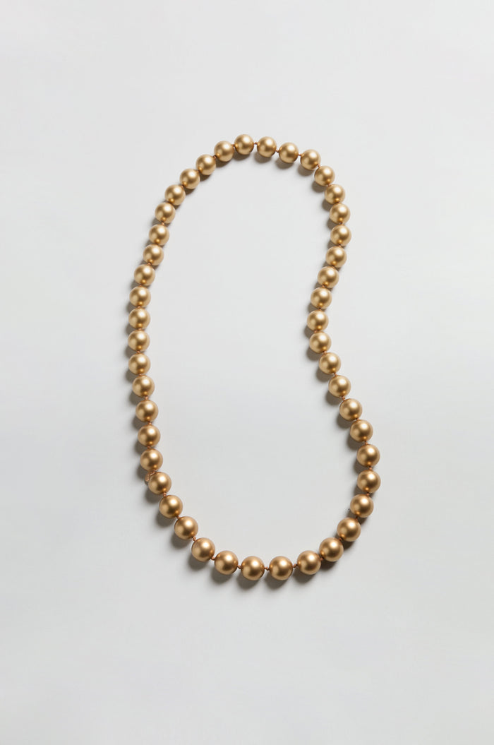 The Long Spheres Necklace is a single strand longline piece with round, gold-colored beads and a stunning gold burnished finish, displayed in an open loop on a simple, light background.
