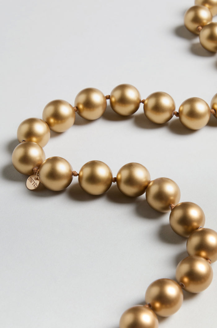 A close-up of the Long Spheres Necklace, showcasing resin spherical beads with a gold burnished finish and a small round tag, elegantly arranged on a light-colored surface.
