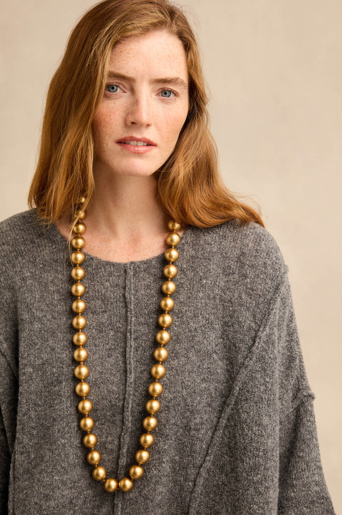 A woman with long auburn hair wearing a grey sweater and the Long Spheres Necklace, which features gold burnished resin spheres, stands against a neutral background.