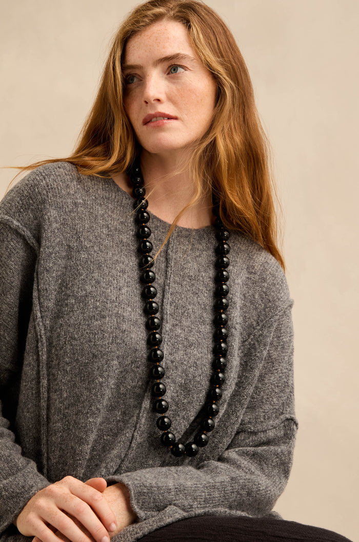 A woman with long auburn hair wears a loose grey sweater and the Long Spheres Necklace, featuring stylish resin spheres, while sitting against a plain beige background.
