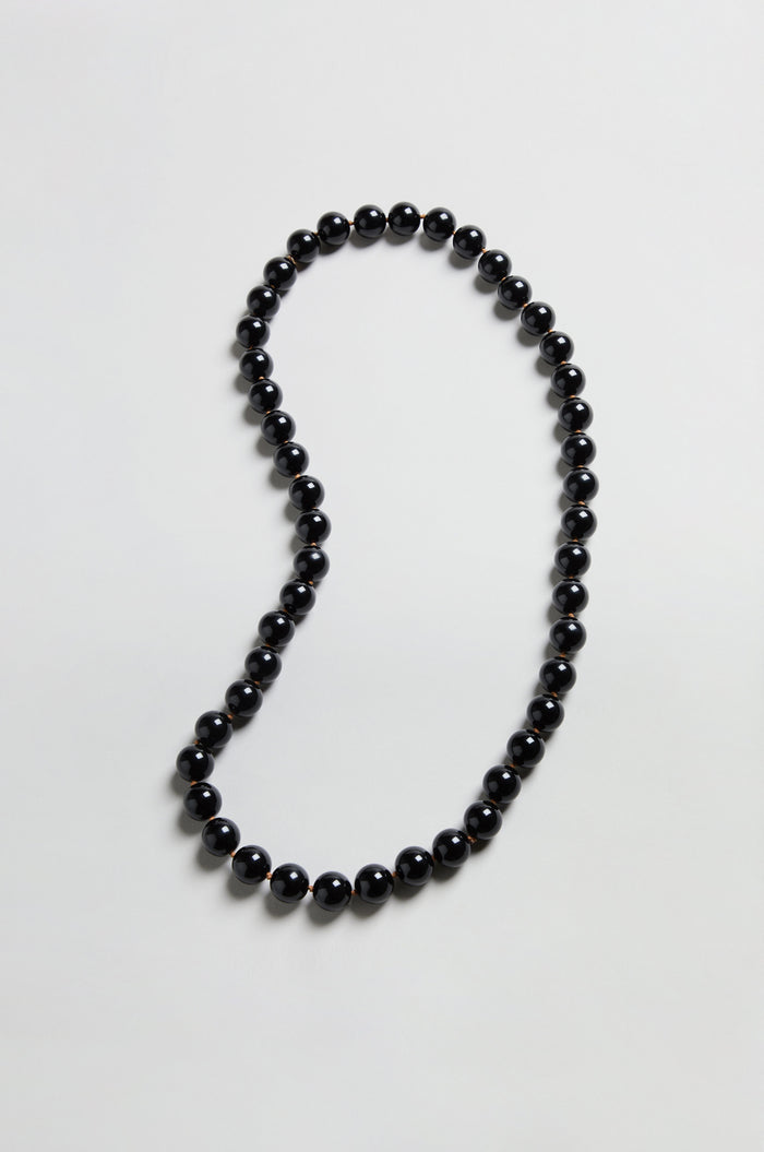 The Long Spheres Necklace features a single strand of round black beads arranged in an irregular loop on a simple, light-colored background.