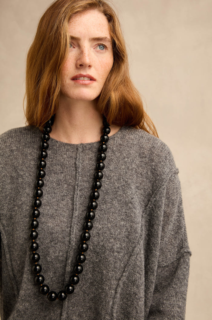 A person with long auburn hair, dressed in a loose grey sweater and wearing the Long Spheres Necklace—featuring striking black resin spheres—gazes slightly to the side against a simple beige backdrop.